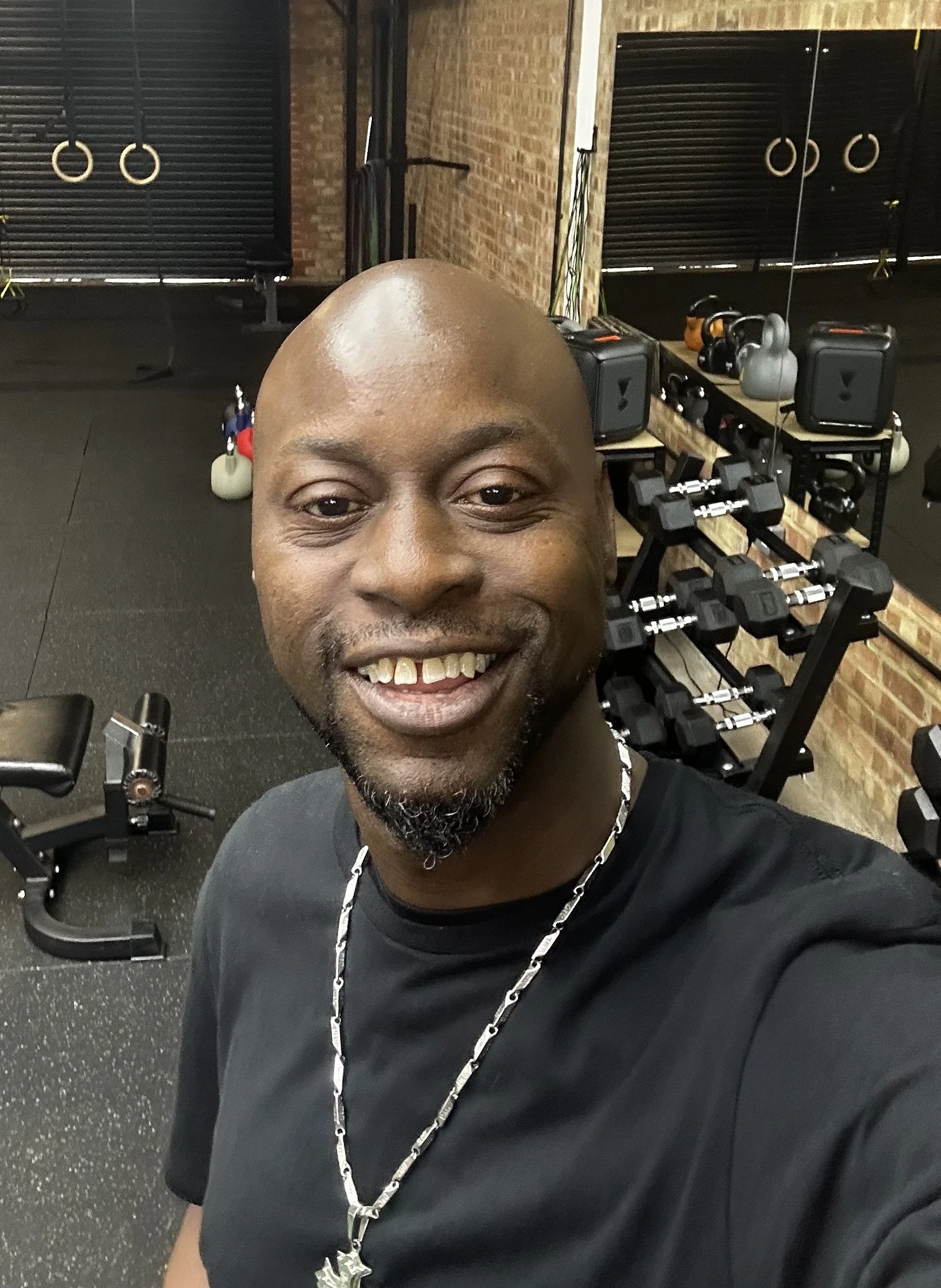 A man taking a selfie in a gym, smiling, with kettlebells, dumbbells, and fitness equipment in the background.
