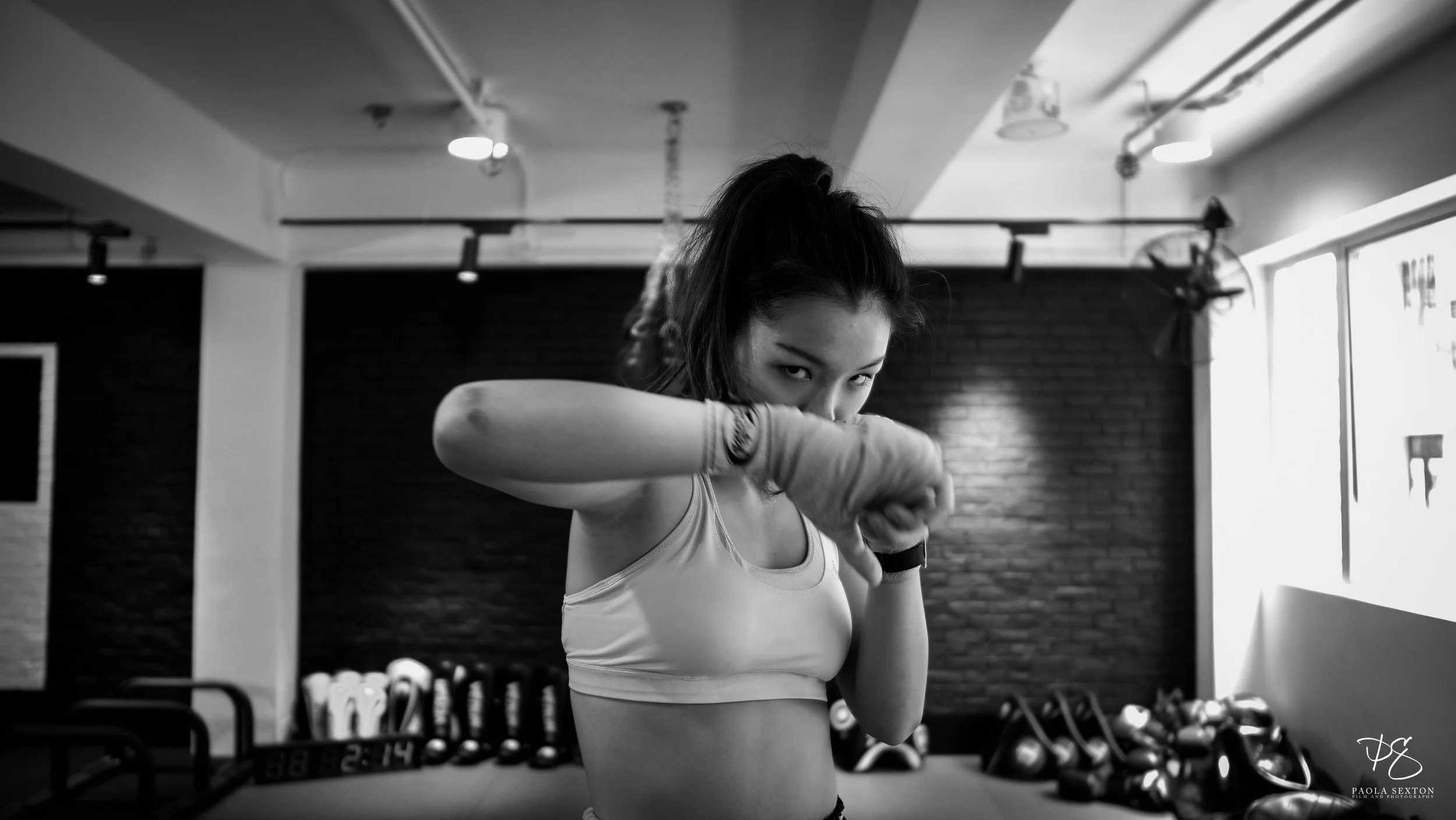 A young woman with dark hair in a high ponytail, wearing a sports bra and boxing gloves, is practicing boxing in a gym. She is focused and looking at the camera while throwing a punch. There are kettlebells and a punching bag in the background.