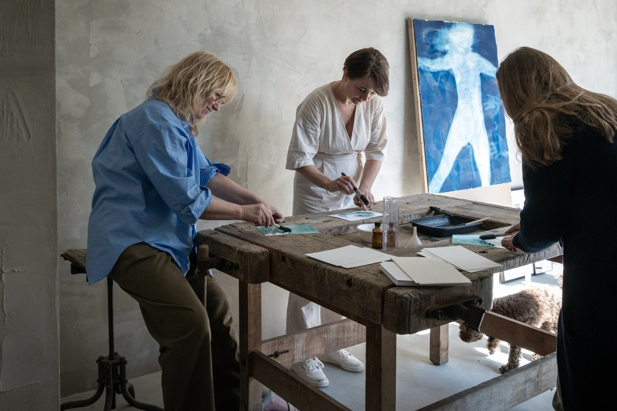 Drie vrouwen werken aan een kunstproject in een studio, met een grote blauwe religieuze afbeelding op de achtergrond, en een hondenras onder de tafel.