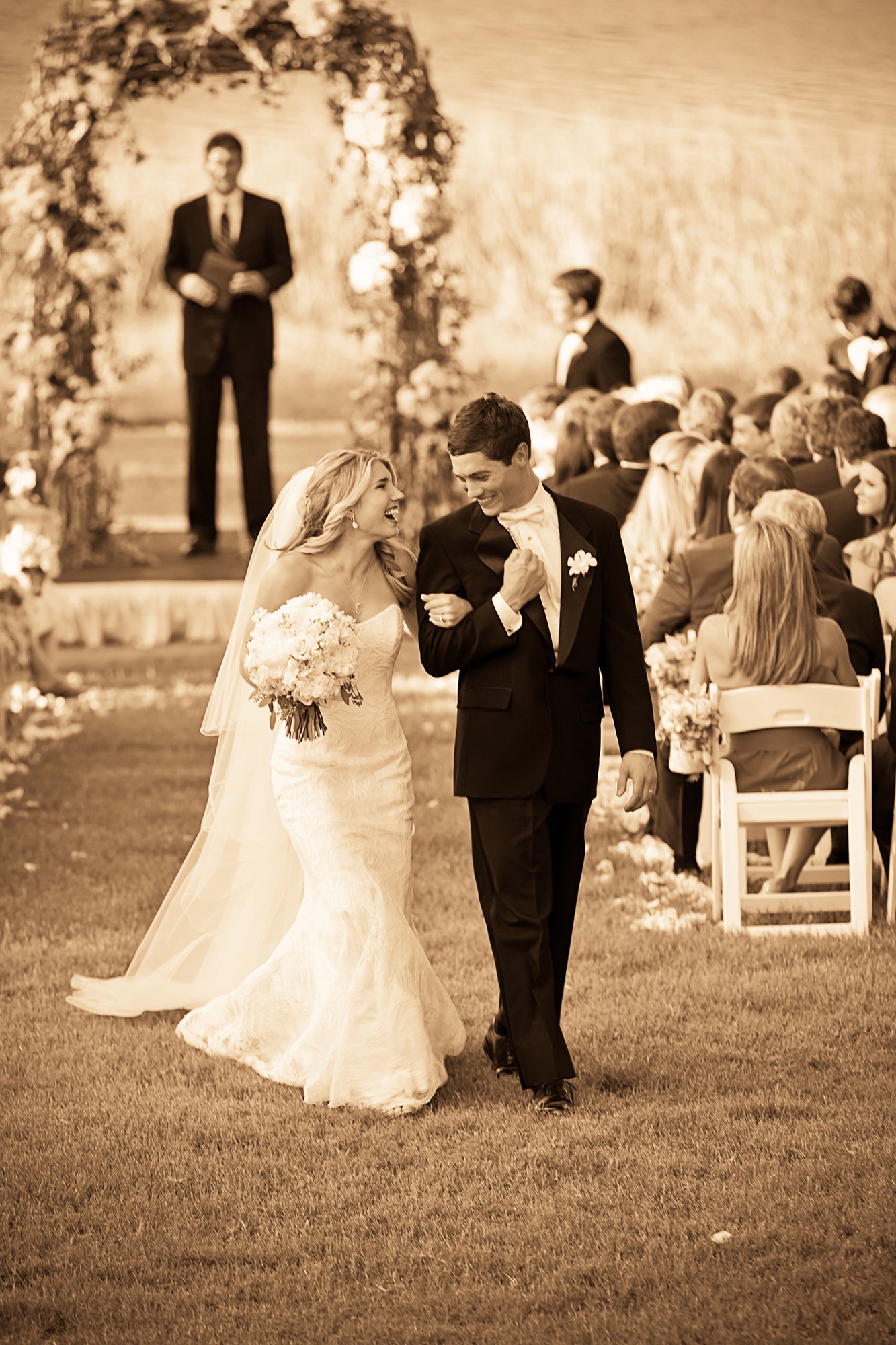 A bride and groom walking down the aisle at their wedding ceremony, surrounded by seated guests, with an officiant standing at a decorated altar in the background.