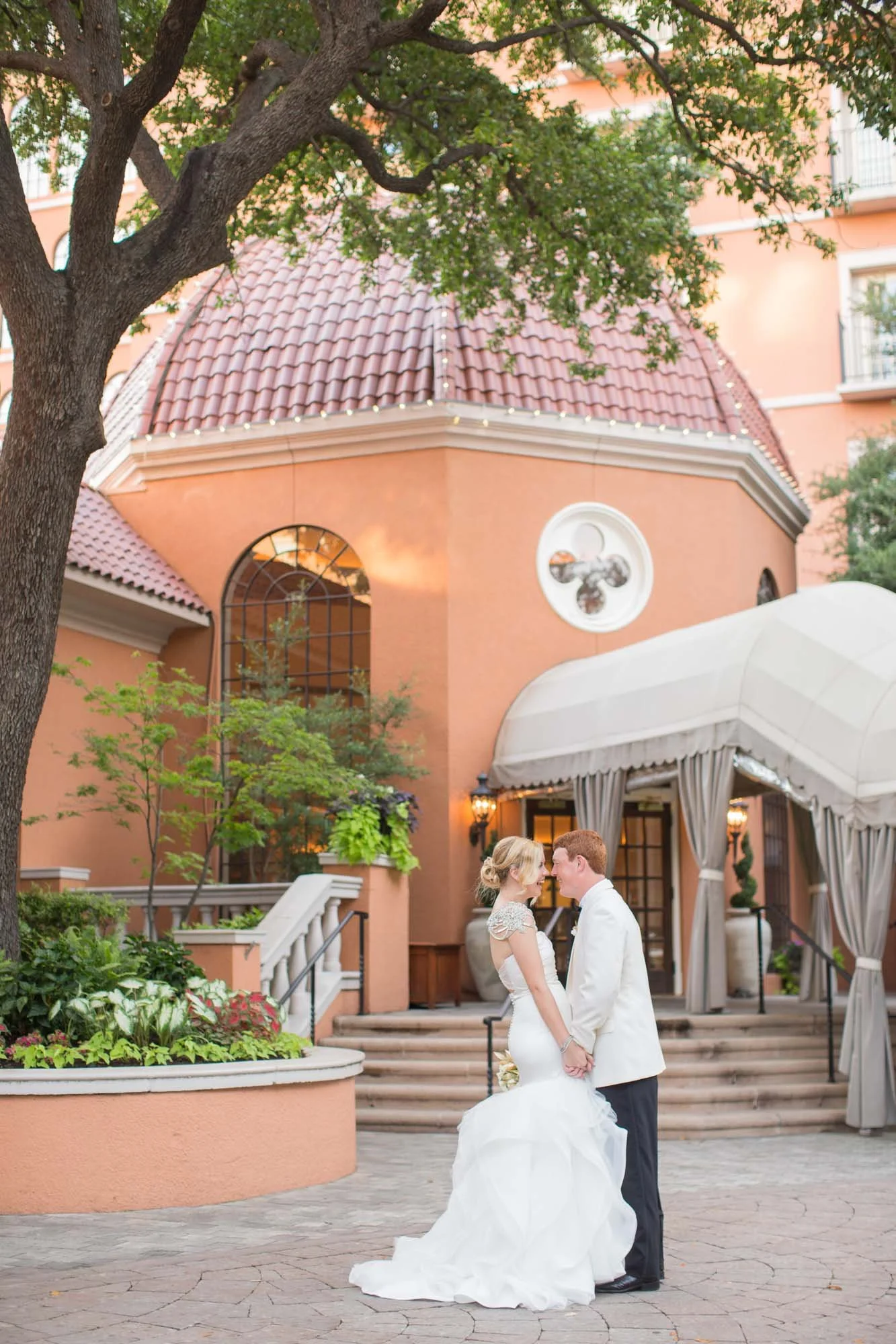 A bride and groom standing face to face outside a peach-colored building with a rounded roof, surrounded by greenery, holding hands and smiling at each other during a wedding.