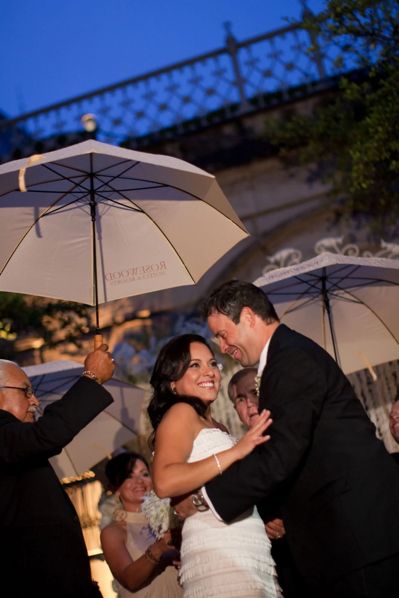 A wedding ceremony outdoors during evening, with the bride and groom smiling at each other under umbrellas held by guests, including older individuals, in front of a decorative wrought iron fence and trees.