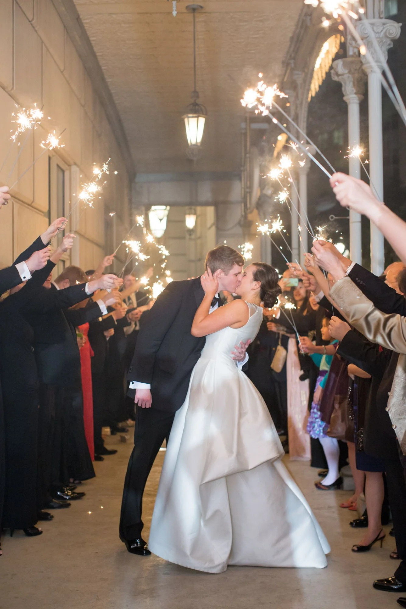 A newlywed couple sharing a kiss under sparklers held by wedding guests during a celebration.
