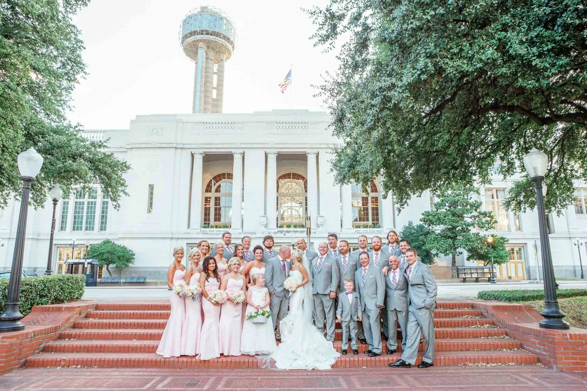 A large group of people dressed in wedding attire, including the bride in a white gown and the groom in a gray suit, standing on red brick steps in front of a white building with tall columns, windows, and clock tower, surrounded by trees and lamppos