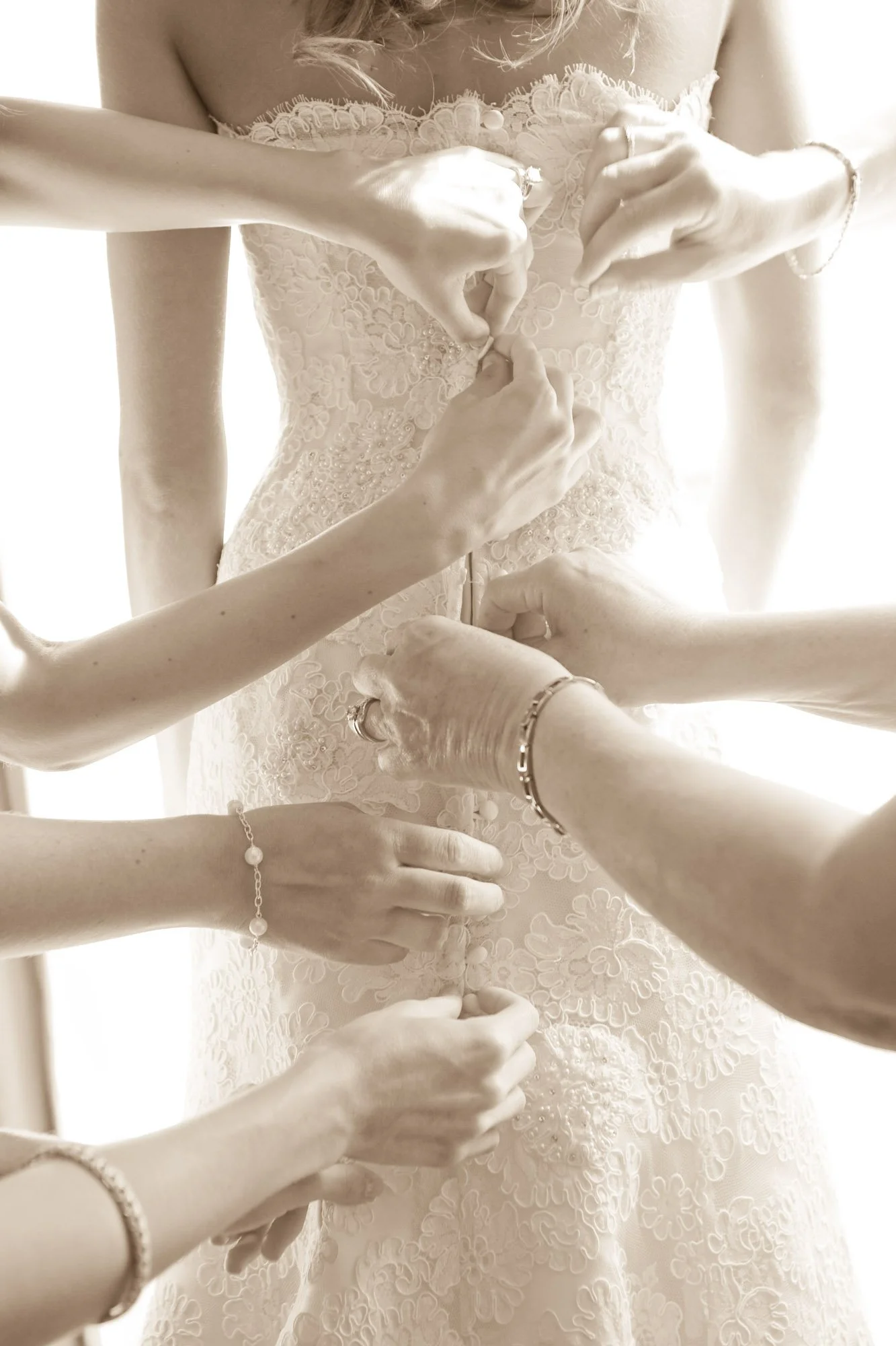 Multiple hands assisting a woman in buttoning up her wedding dress, close-up shot in sepia tone.
