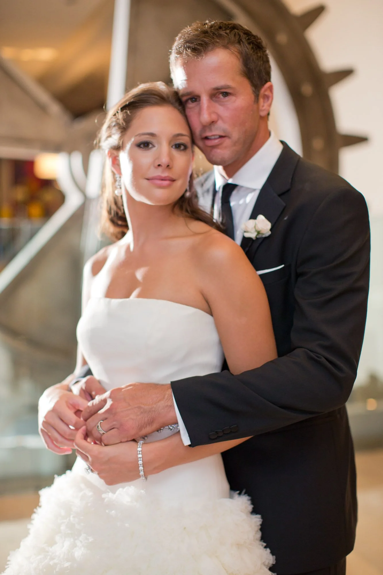 A bride and groom on their wedding day, standing close together indoors. The bride is wearing a strapless white wedding gown with a textured skirt and is accessorized with earrings, a bracelet, and a ring. The groom is dressed in a black suit with a 