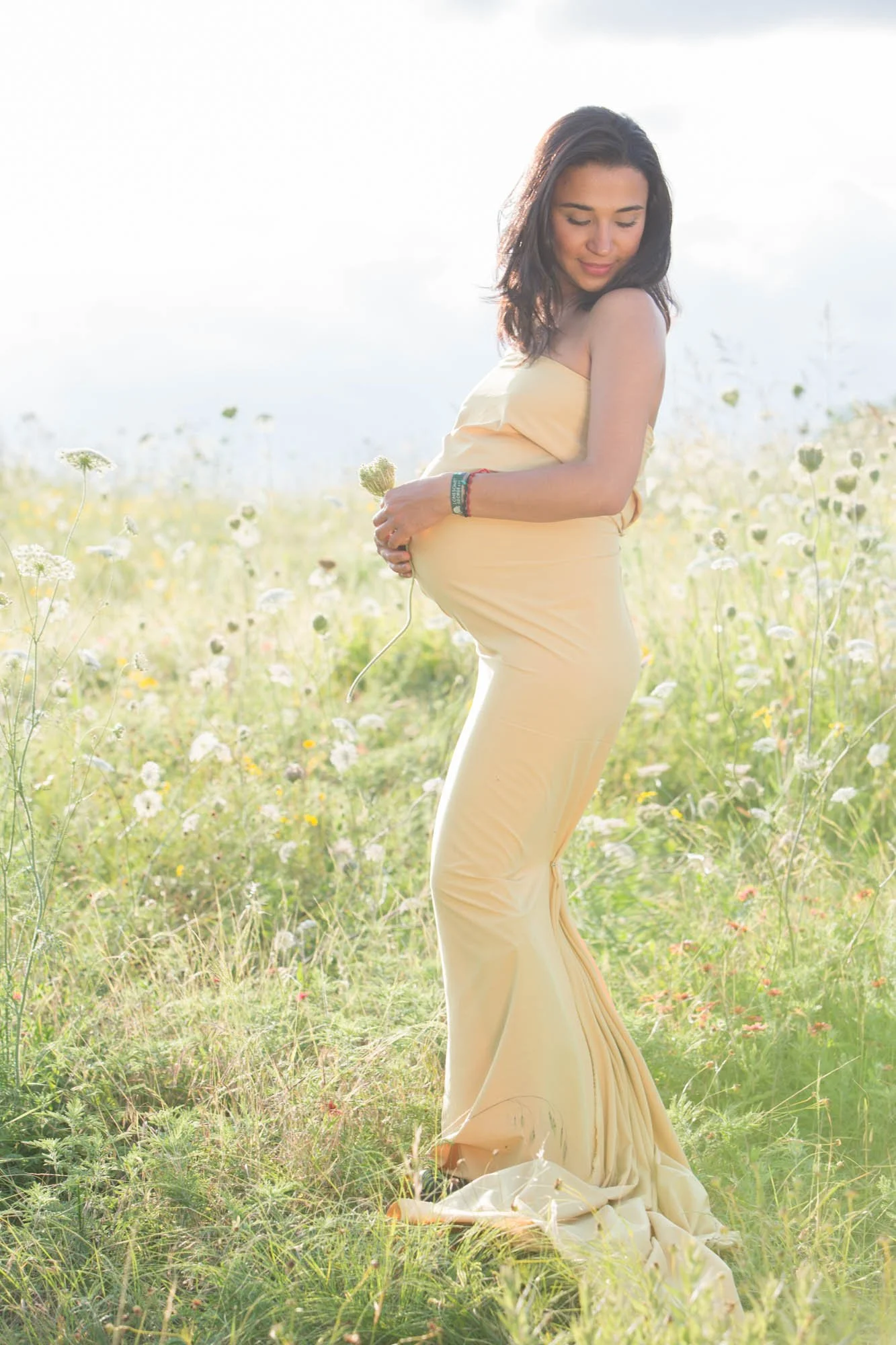 A pregnant woman wearing a yellow dress is standing in a field of wildflowers during daylight, holding a flower near her belly and smiling softly.