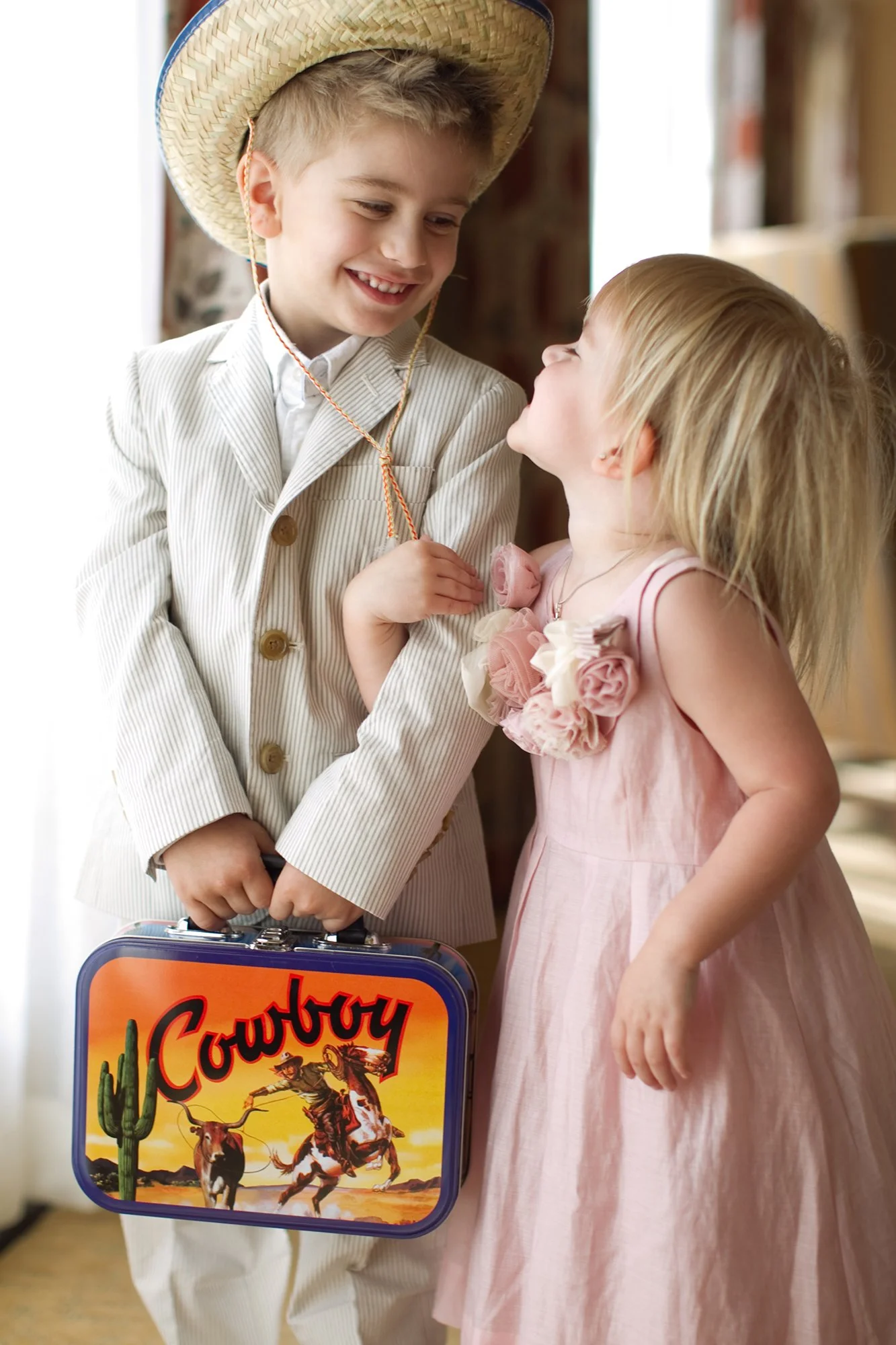 A boy dressed as a cowboy and a girl in a pink dress sharing a joyful moment indoors. The boy wears a cowboy hat and a striped suit, holding a lunchbox with a cowboy graphic, and the girl looks up at him with a smile.