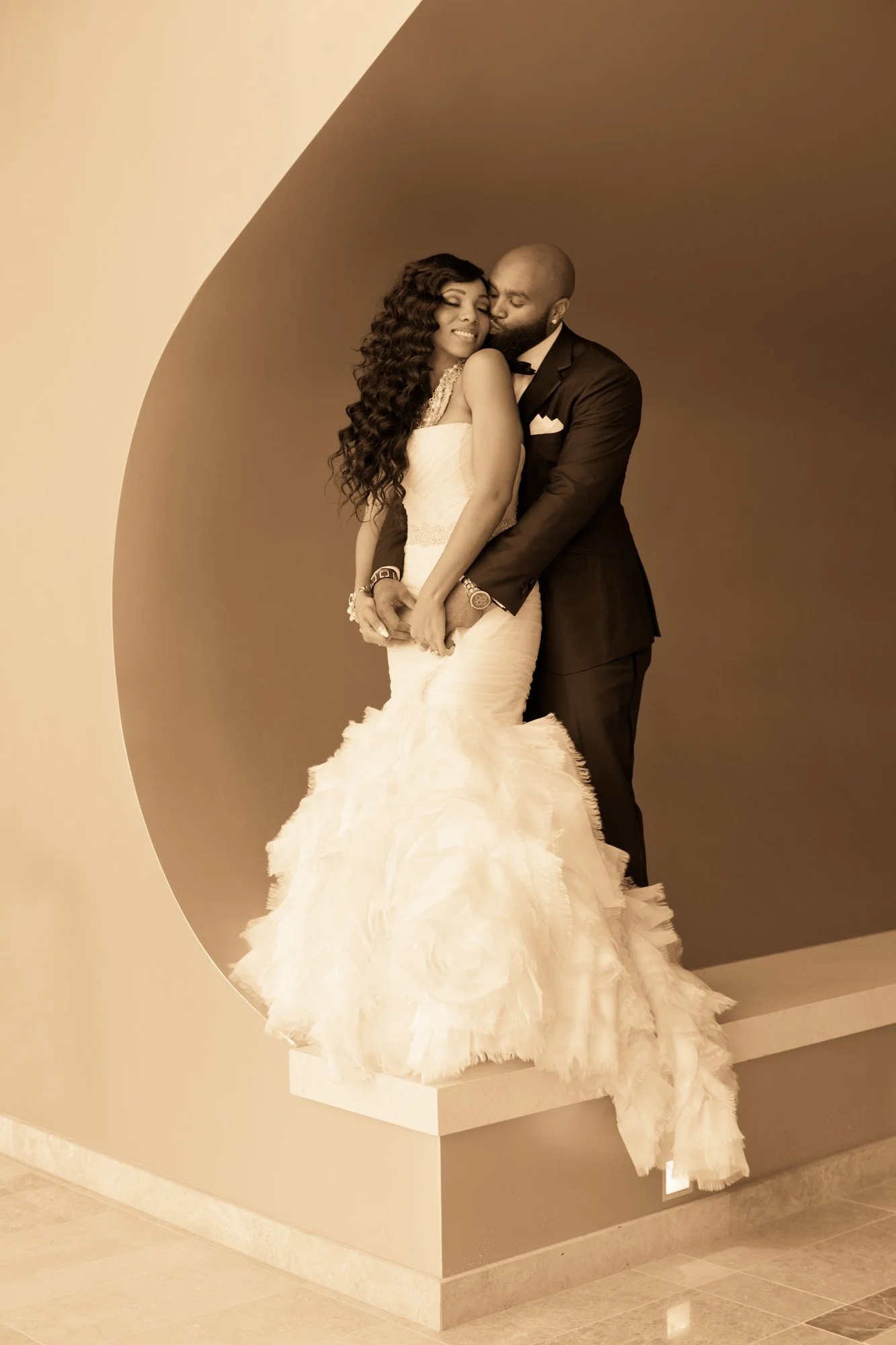 A bride and groom in formal attire are embracing each other. The bride is wearing a white wedding gown with a ruffled train and the groom is in a black suit with a bow tie. They are standing on a platform against a dual-colored background.