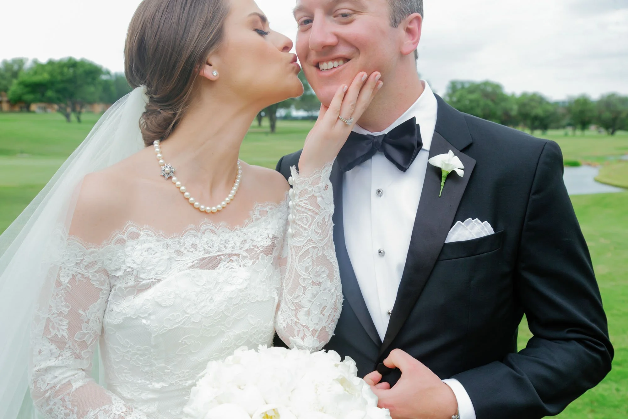 A bride and groom in wedding attire sharing a kiss on a golf course with green grass and trees in the background.