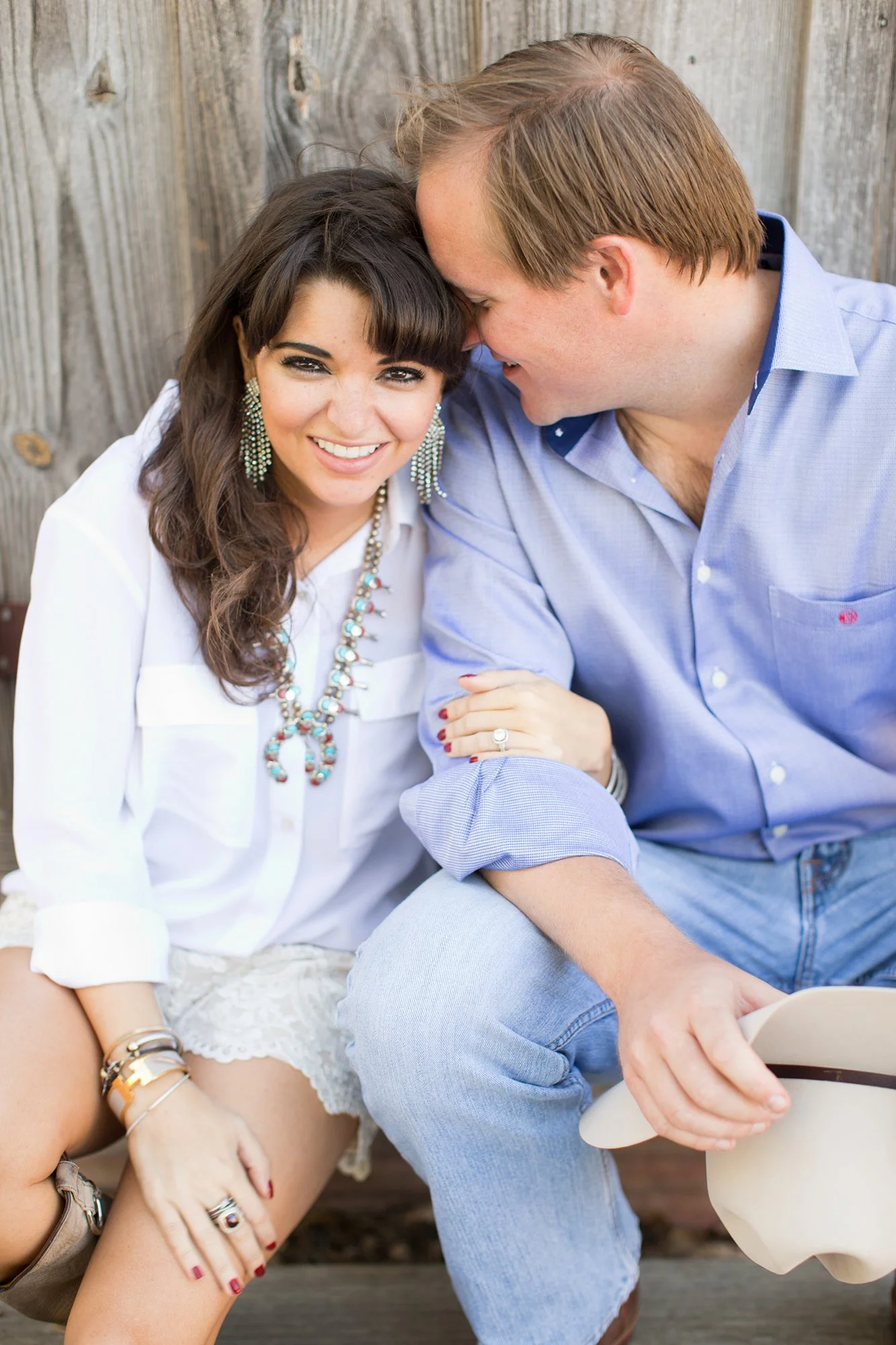 A smiling woman and a man sitting close together outdoors, leaning their foreheads touching. The woman has dark hair, wearing a white blouse, colorful jewelry, and has a ring on her finger. The man has light brown hair, wearing a light blue shirt, an