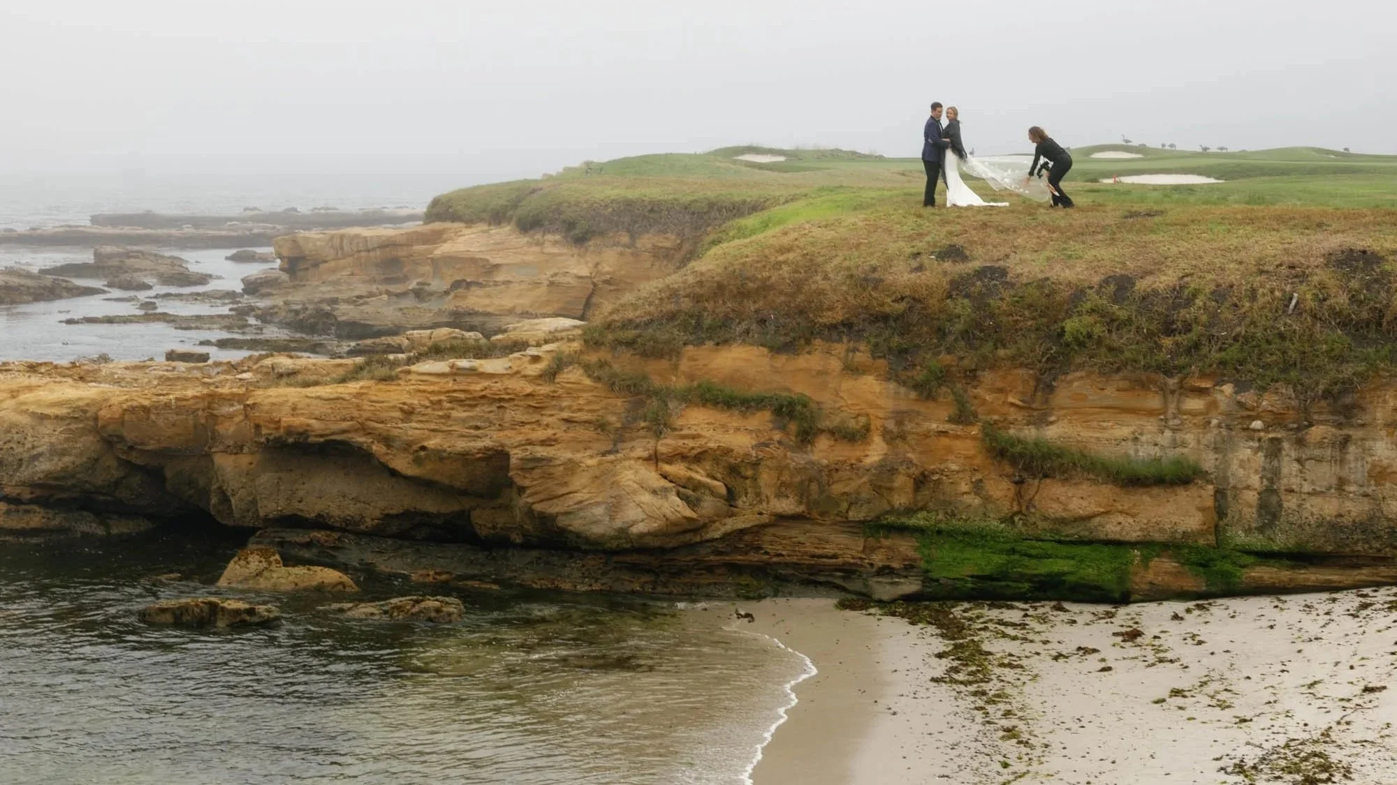A wedding ceremony taking place on a grassy cliff overlooking the ocean, with a bride and groom holding hands and a person adjusting the wedding dress or veil.