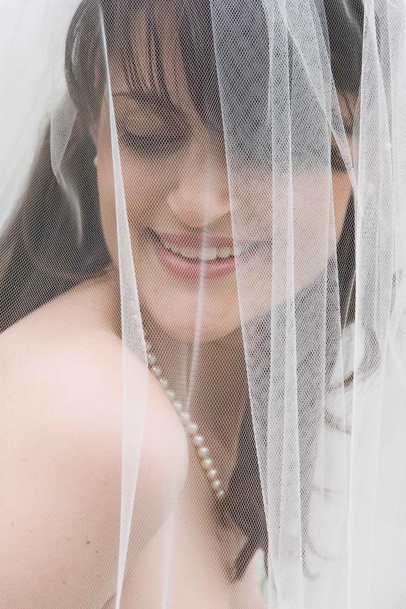 Close-up of a smiling bride with dark hair and a pearl necklace, partially covered by a white veil.
