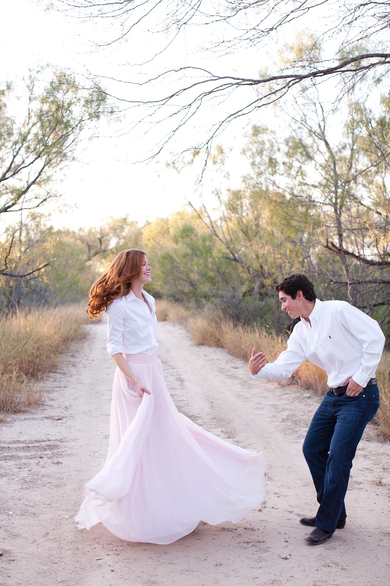 A young couple dancing on a dirt path surrounded by trees with green and dried foliage, during sunset, with the woman in a long pink skirt and white blouse and the man in dark jeans and a white shirt.
