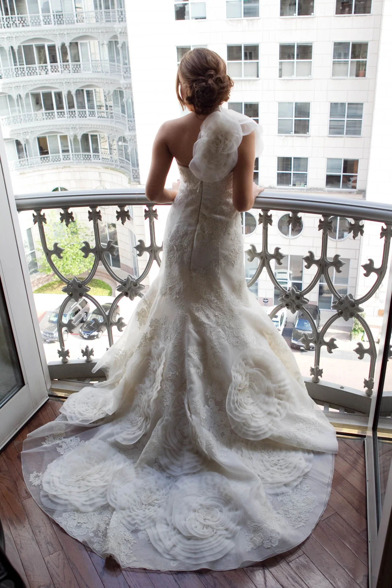 A woman in a wedding dress stands on a balcony overlooking a city street, with her back facing the camera.