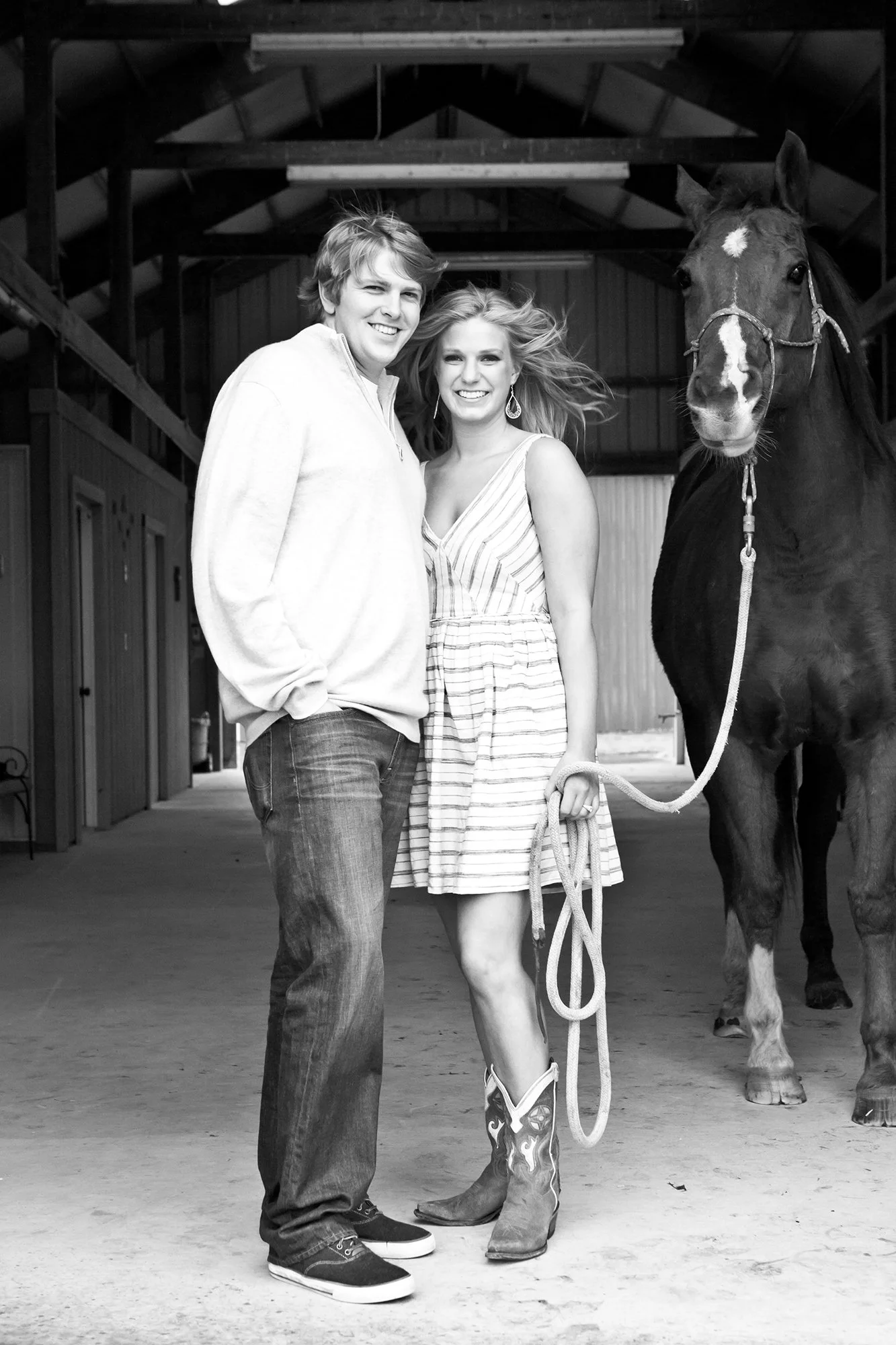 A black-and-white photo of a man and woman standing inside a barn, smiling at the camera, with a horse on the woman’s right side.
