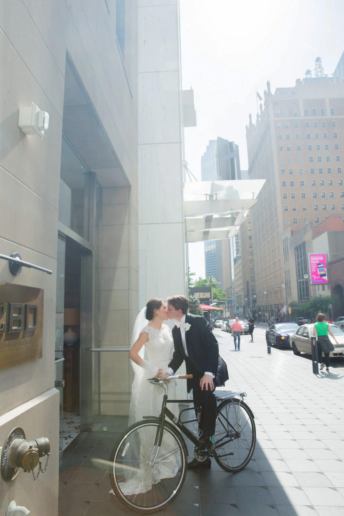 A newlywed couple kissing outside a building on a city street, with the groom on a bicycle and the bride in a white wedding dress.