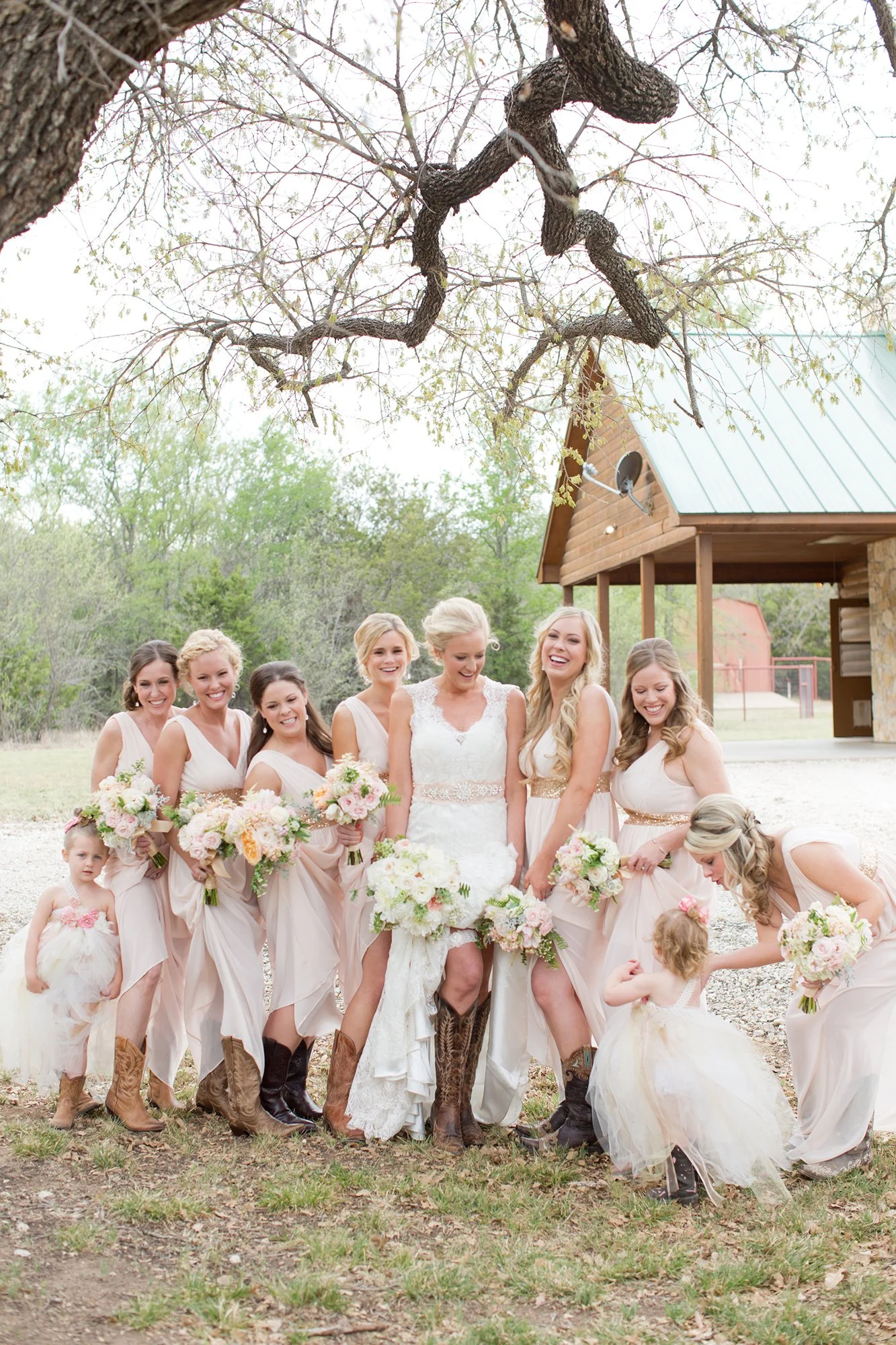 A bride and six bridesmaids with two flower girls outdoors, holding bouquets, in front of a rustic barn with a metal roof, under a sprawling tree with twisting branches.
