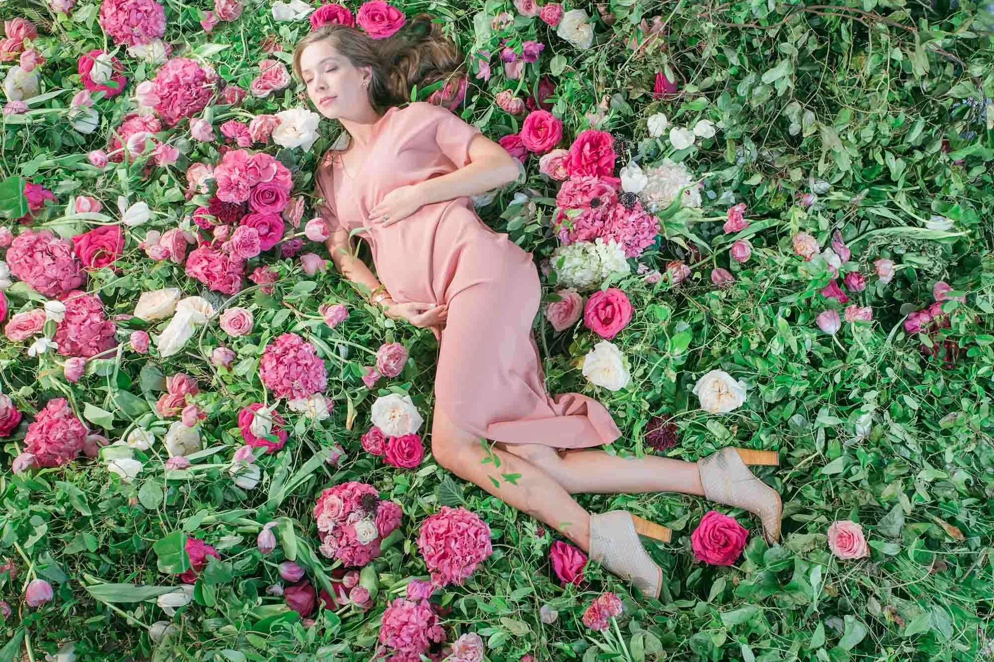 A woman in a pink dress lying on a bed of pink, white, and purple flowers with closed eyes.