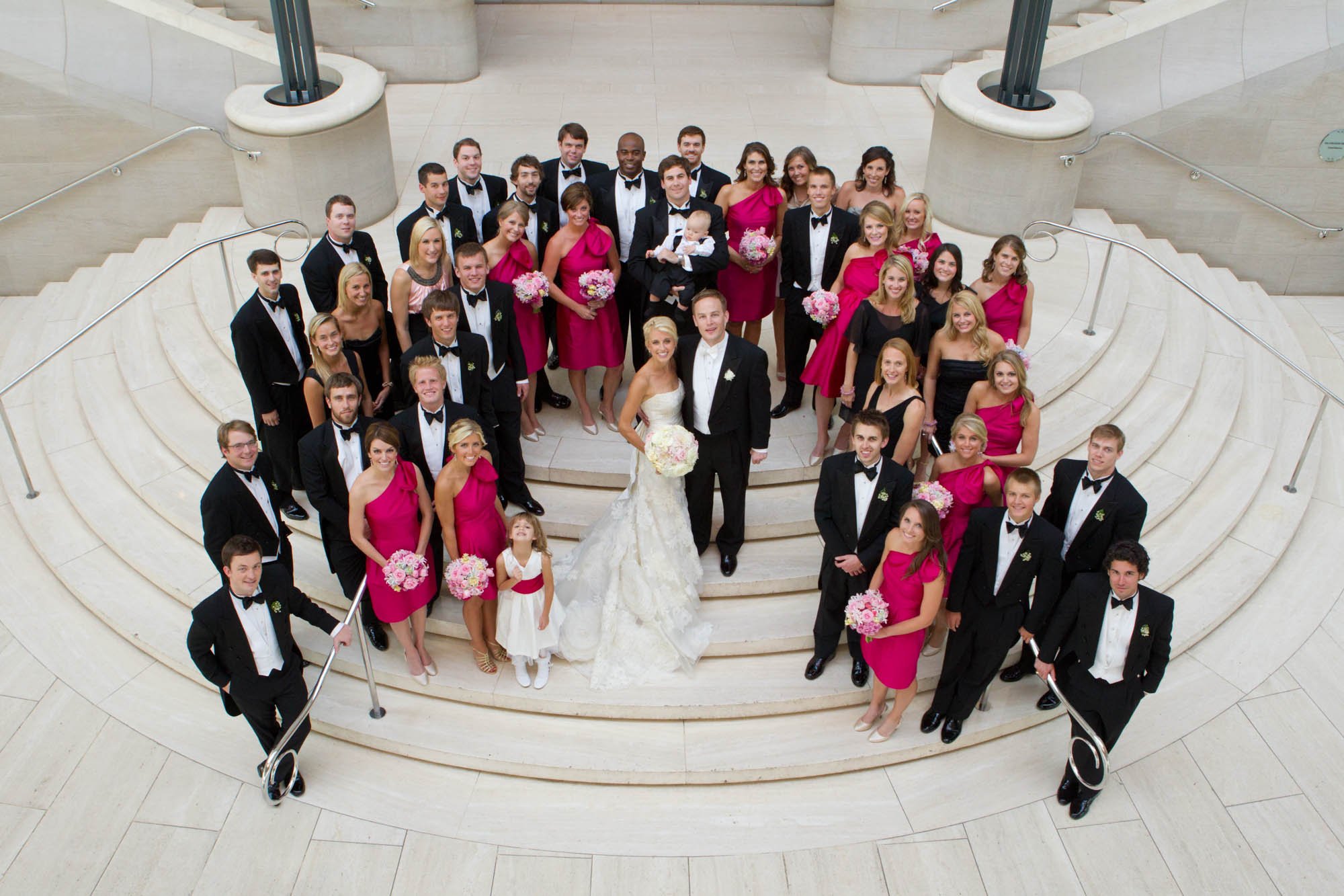 A wedding party on a staircase with the bride and groom in the center, surrounded by bridesmaids in pink dresses and groomsmen in black tuxedos, all smiling for a group photo.