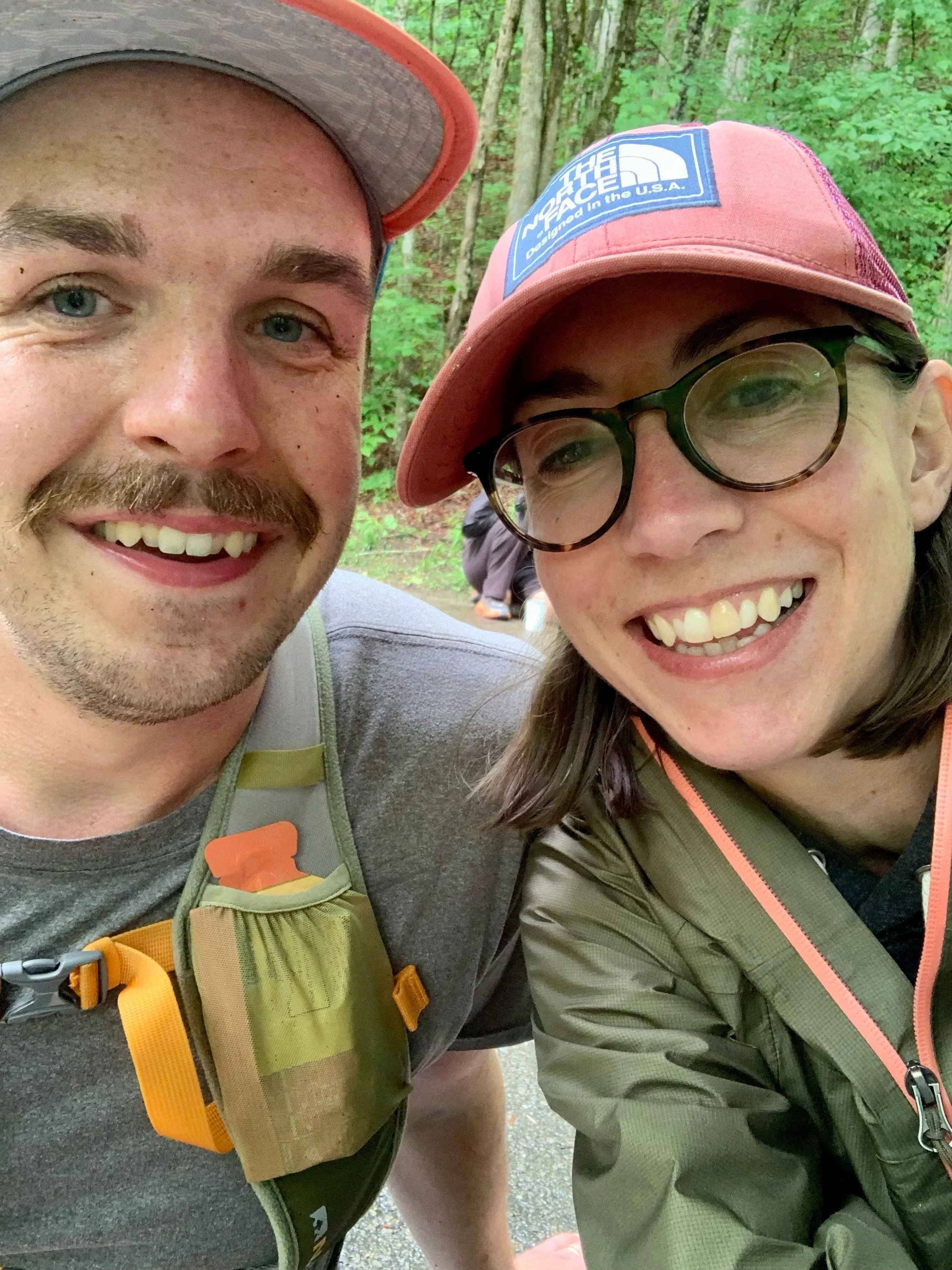Close-up of two smiling individuals, a man and a woman, taking a selfie outdoors in a forest setting. The man has a mustache, beard, and is wearing a gray cap and a hydration pack. The woman is wearing a pink cap with The North Face logo, glasses, and a green jacket. Green foliage and trees are visible in the background.