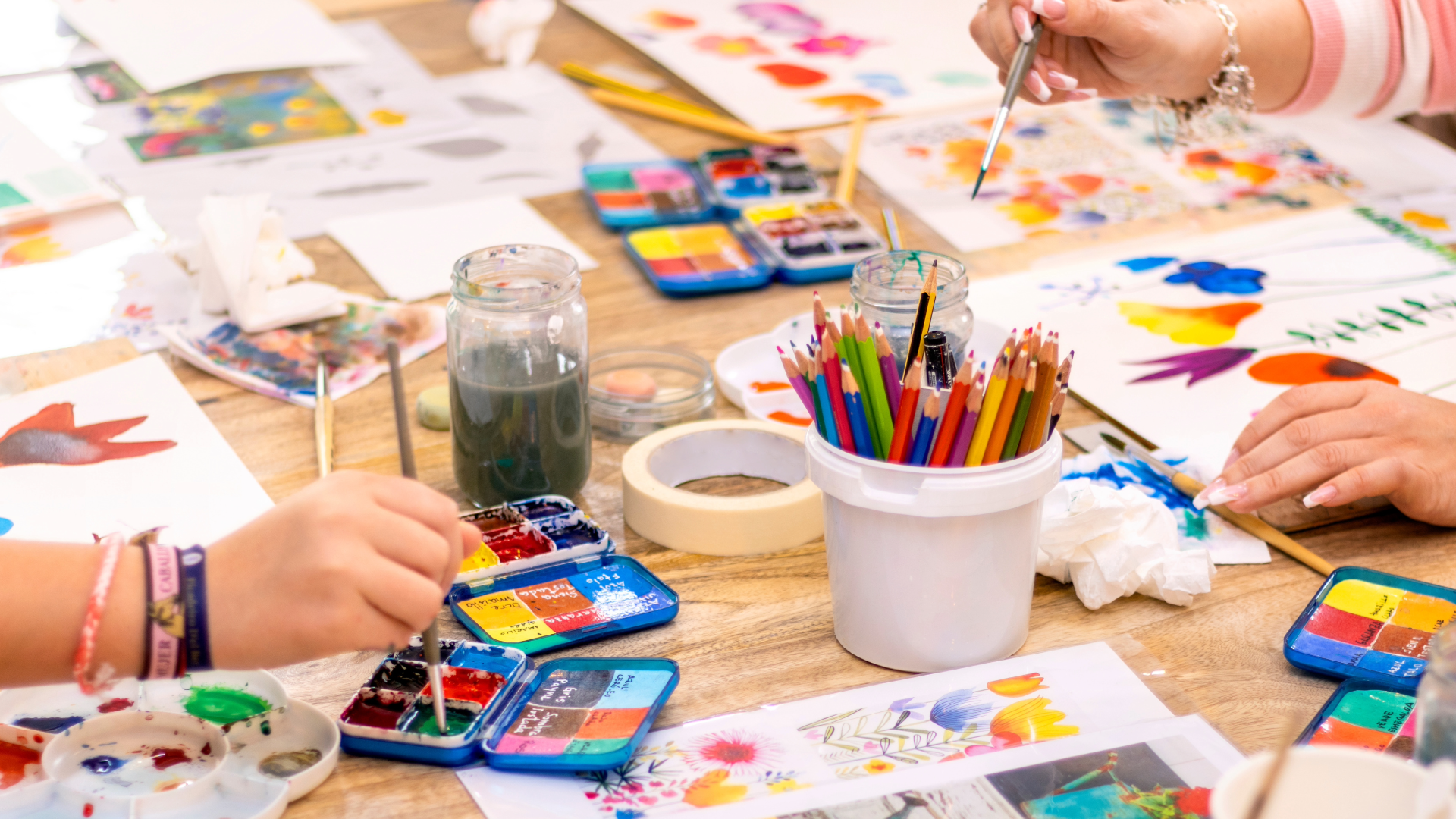 People creating colorful watercolor paintings with paint, brushes, and colored pencils on a wooden table.