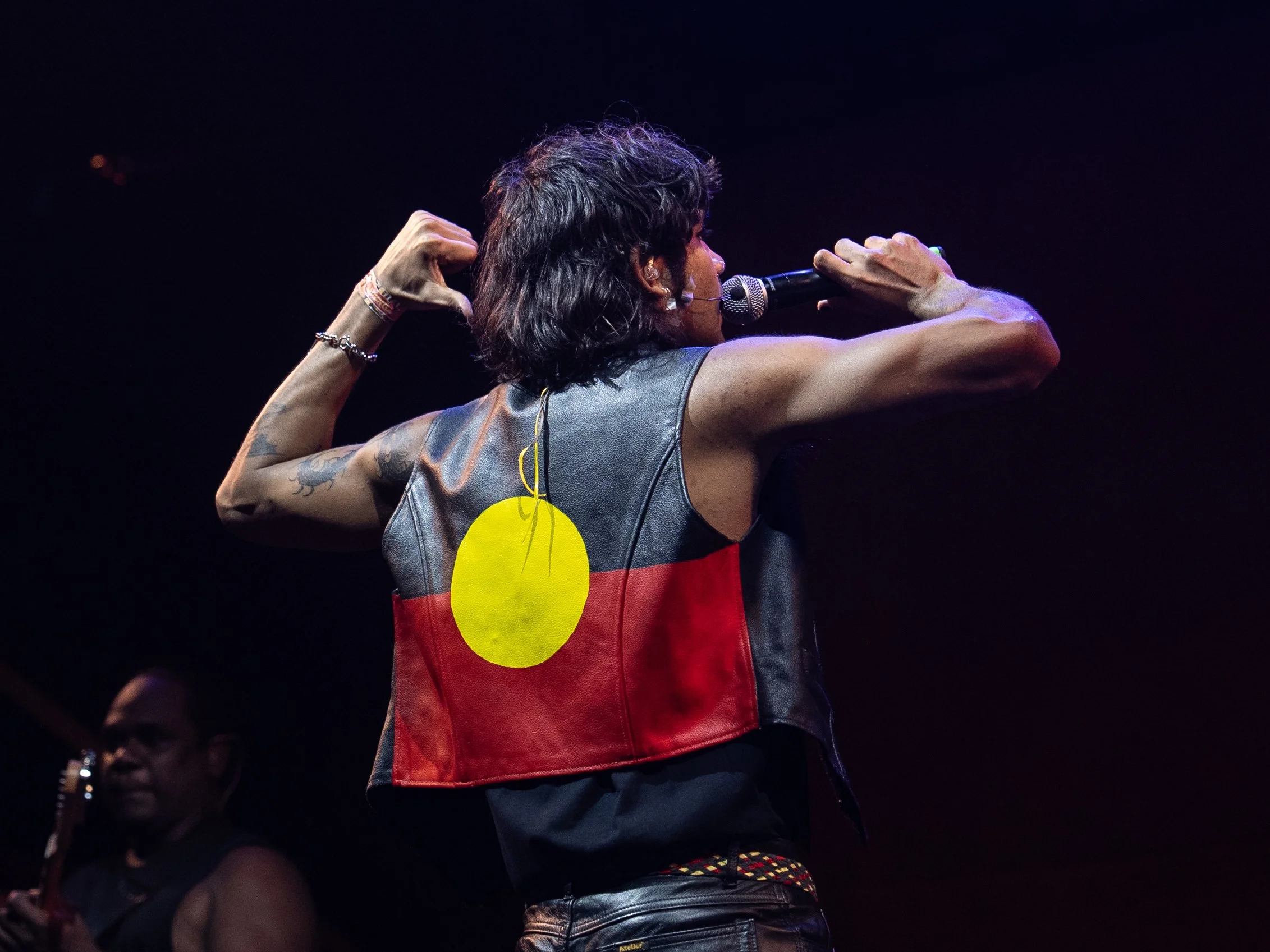 An Indigenous man holding a microphone with both arms up. He is wearing a black leather vest with the Aboriginal flag emblazoned on the back.
