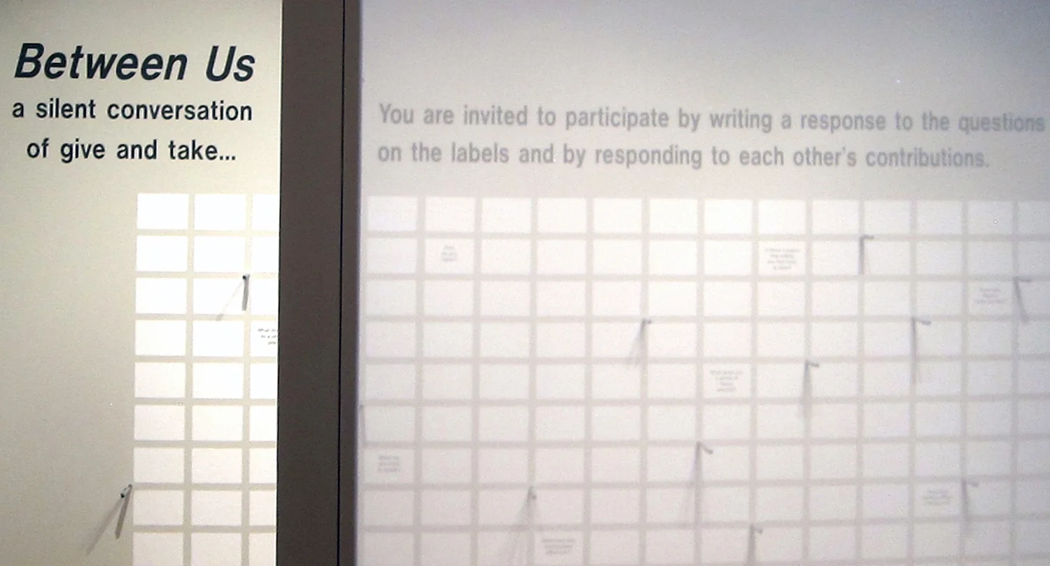 A wall text invited viewers to respond to 11 open-ended questions that were printed and distributed throughout a grid of blank labels. Participants could stand at the wall to write on one of the blank labels surrounding the questions. Writing pens were mounted in metal sleeves placed in the wall surface. A custom bench designed with slots for notebooks and pens provided an alternative seat for response or reflection.