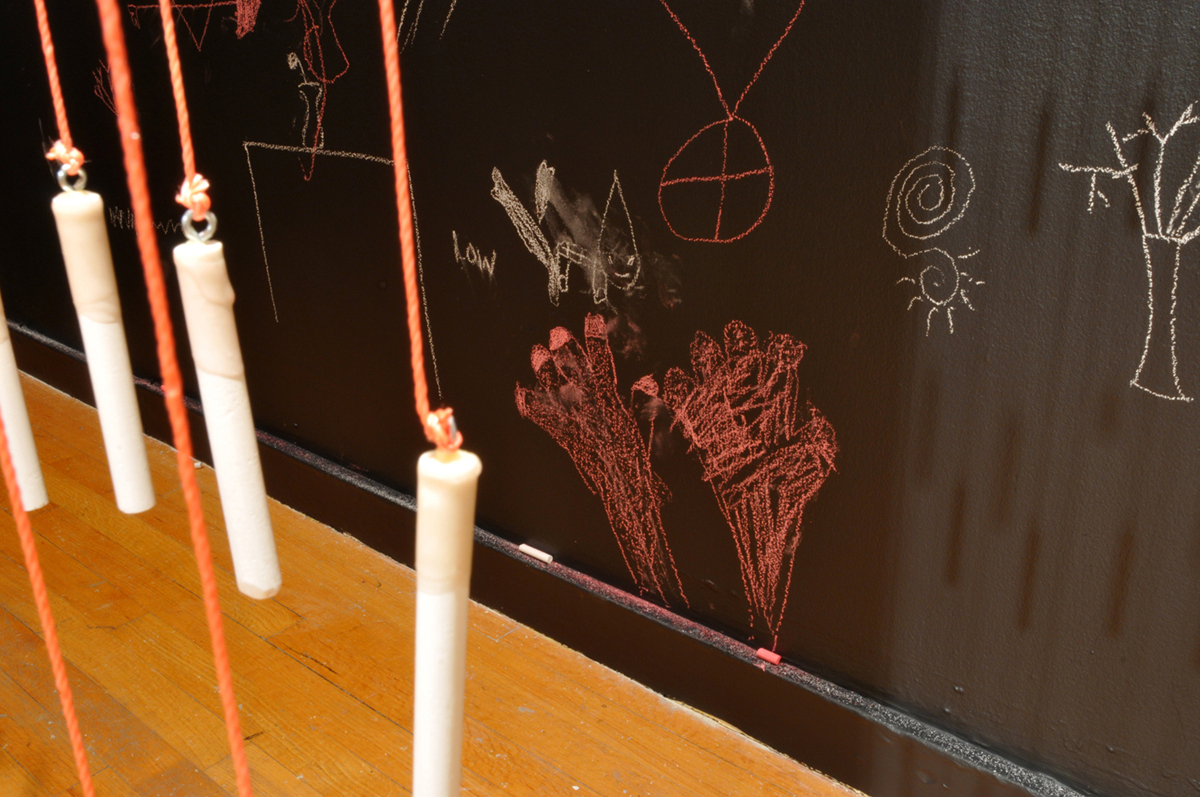 Hundreds of pieces of chalk suspended on shimmering nylon cords formed the central core of chalk talk . Tiered layers of weighted string defined a fluid boundary of passage and an interactive writing system surrounded by black walls. Viewers quickly understood that they could use the stretchy chalk lines and blackboard surfaces to join an on-going conversation. Nothing was erased, rather like graffiti, walls filled with overlapping comments, responses, drawings, marks and gestures. Chalk lines tangled and were untangled/ tended by participants as they used the interactive space.