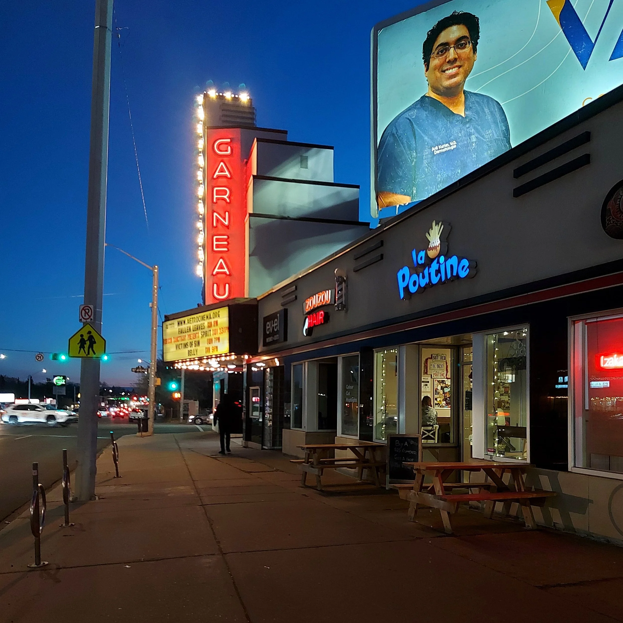 Night scene outside a restaurant called La Poutine with outdoor seating, a bright, glowing, vertical sign saying Garneau. The Historic Garneau Theatre Building.