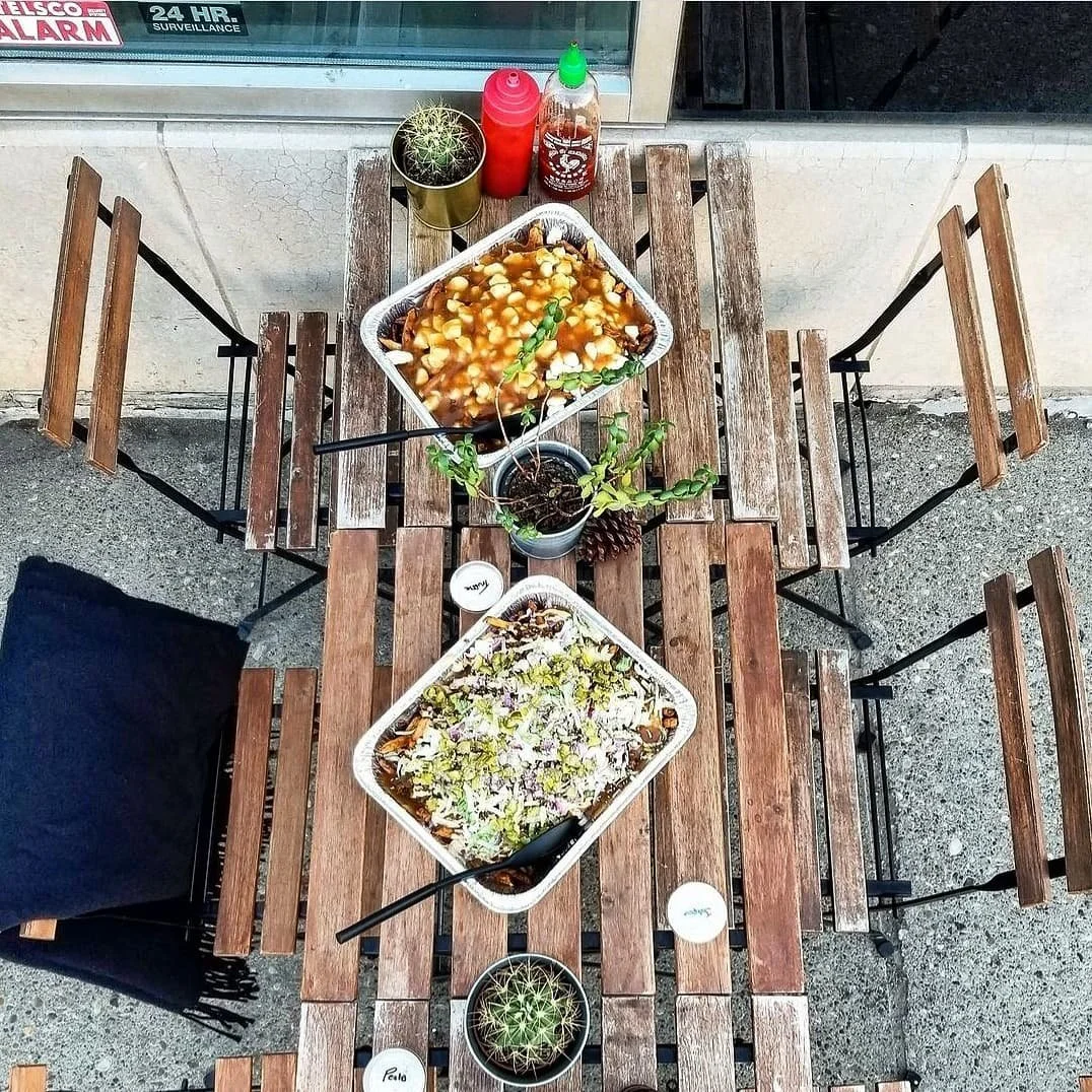 Outdoor wooden table with two aluminum trays of food, assorted condiments, potted cacti, and small candles.
