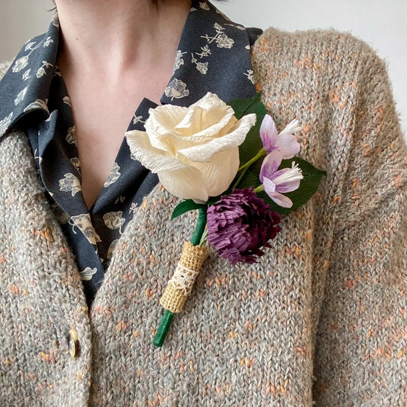 Handmade paper flower boutonniere with a cream rose, purple blooms, green leaves, and a burlap-wrapped stem