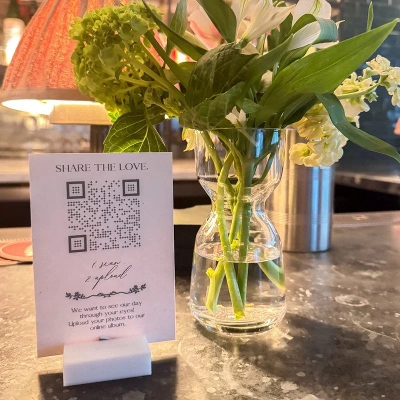 Wedding photo sharing QR code sign displayed beside a vase of fresh flowers
