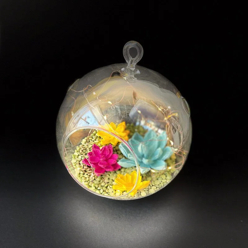 Colorful paper flowers and greenery with fairy lights arranged inside a clear hanging glass terrarium