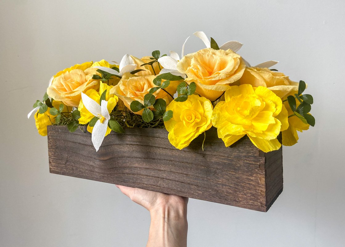 Yellow and peach paper roses with white flowers and greenery arranged in a rustic wooden box