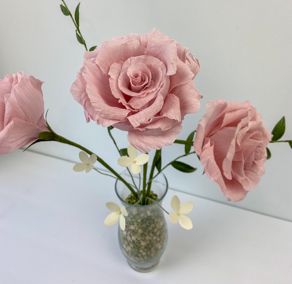 Pink paper roses with delicate white flowers and greenery arranged in a small glass vase