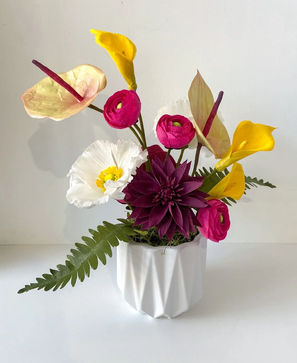 Colorful paper flower arrangement with yellow calla lilies, pink ranunculus, white poppy, burgundy dahlia, and greenery in a white vase