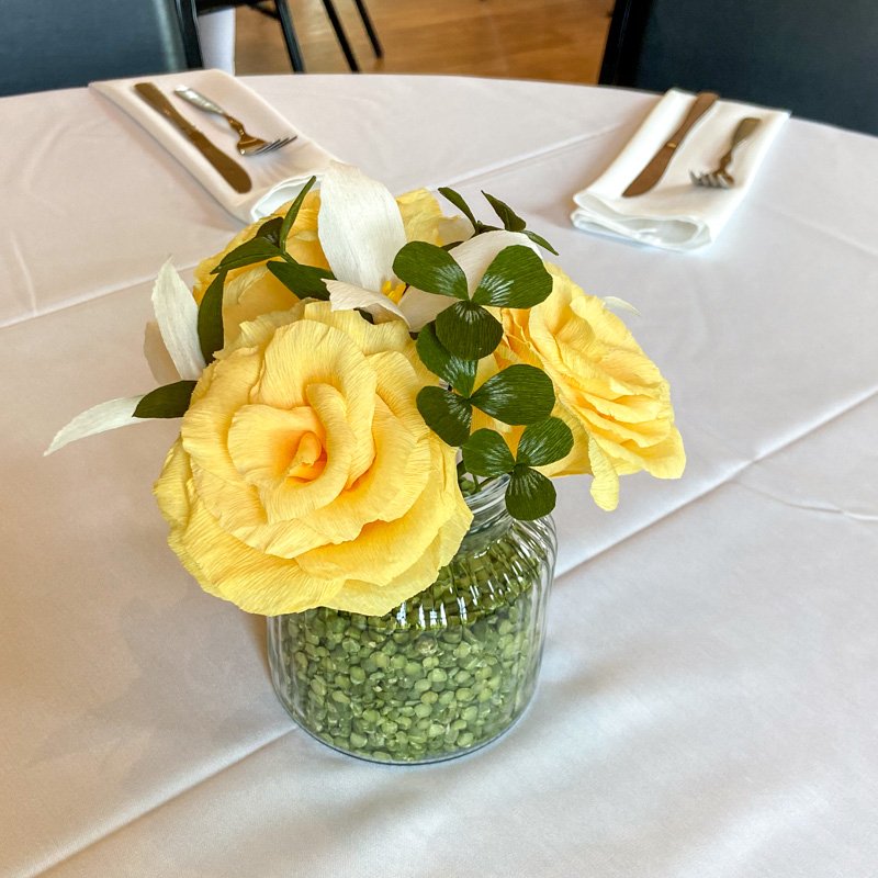Yellow paper roses with white flowers and green leaves arranged in a glass vase on a table