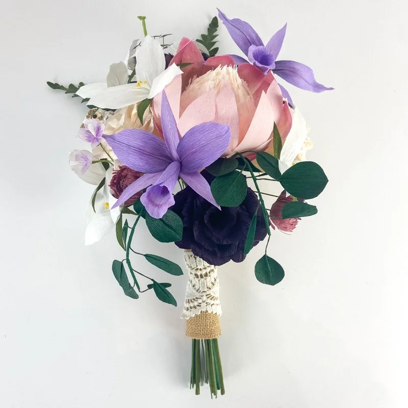 Handmade paper wedding bouquet with purple orchids, blush protea, white flowers, dark purple blooms, and cascading greenery