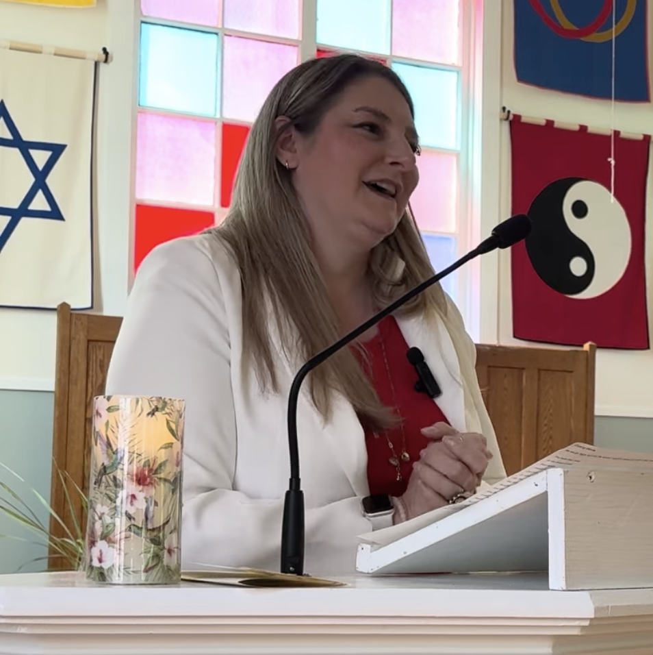 White woman in a red shirt and white blazer giving a speech against a stained glass window