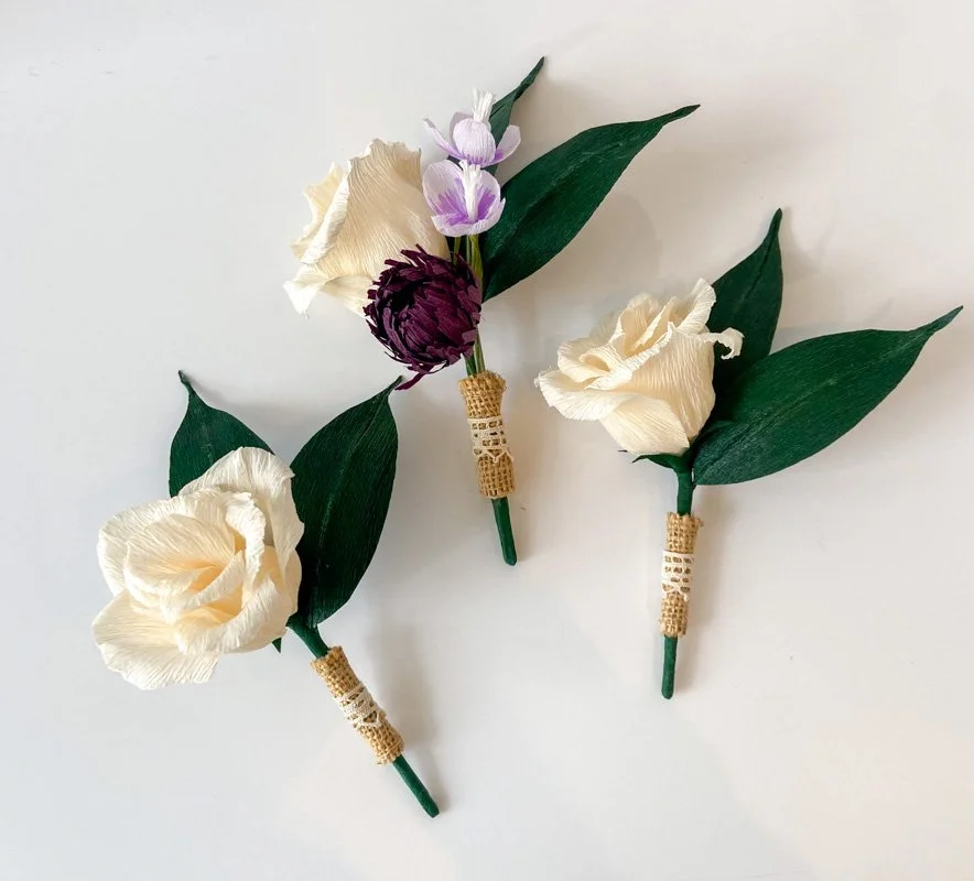 Handmade cream paper flower boutonnieres with green leaves, burgundy and lavender accents, and vintage lace-wrapped stems