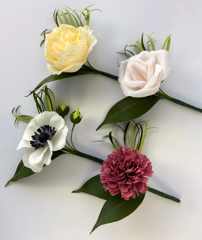Four handmade paper flower boutonnieres featuring yellow, blush, white, and burgundy blooms with green foliage