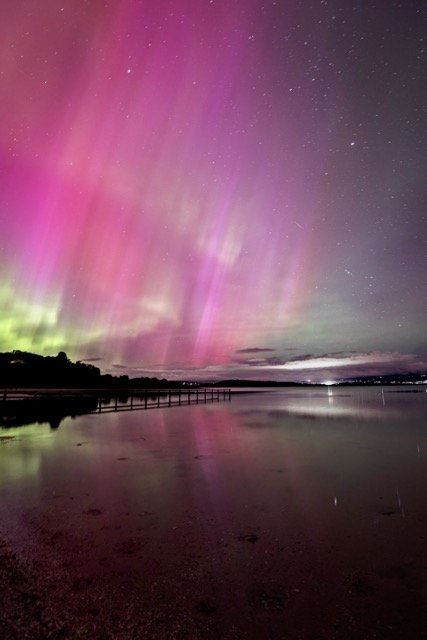 	
Southern lights over a body of water with a shoreline and a pier, starry night sky.