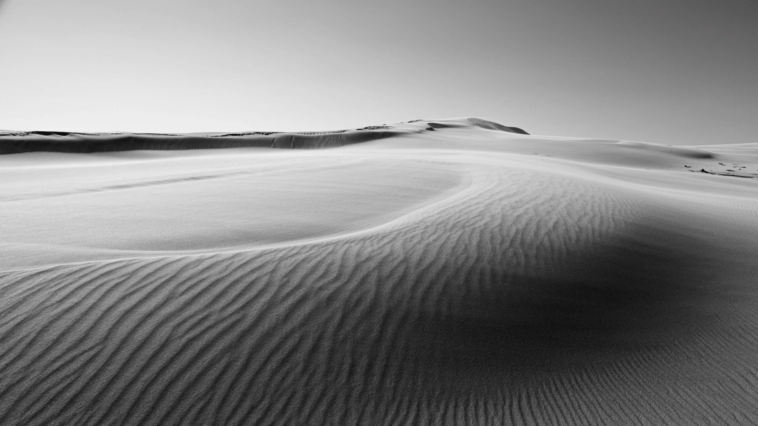 Black and white photograph of sand dunes with textured ripples and smooth curves, under a clear sky.