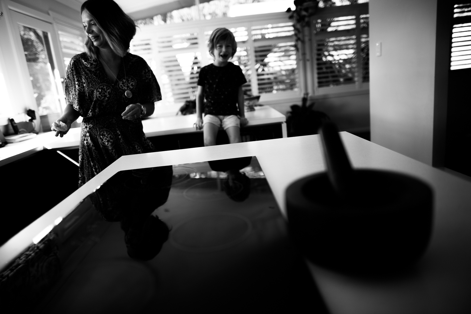 A fun family photo during a lifestyle session in Wollongong. A boy sits on the bench as his Mum darts around the kicthen. Captured by Cindy Cavanagh, a Sydney lifestyle photographer.