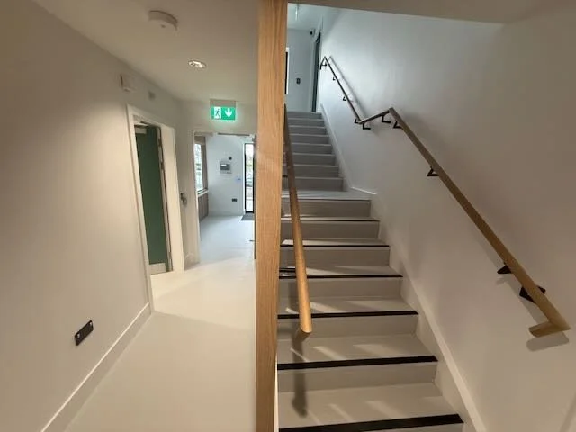 Interior view of a modern building with a staircase on the right side and a hallway on the left, featuring white walls, a wooden handrail, and emergency exit signs.