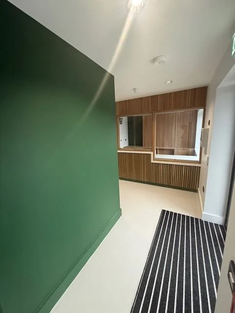 Interior of a modern building with a green wall, wooden reception desk, and black striped floor mat.