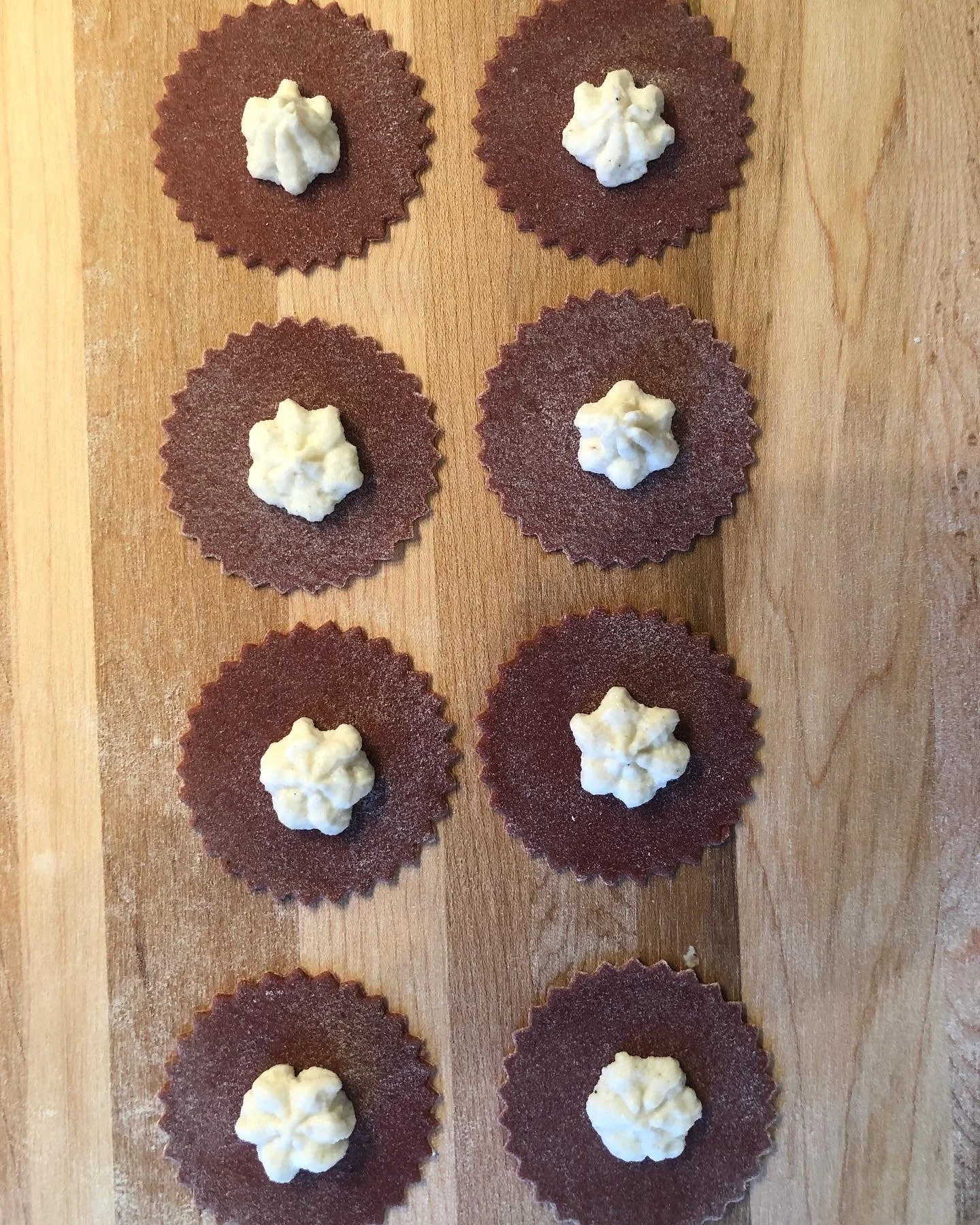 Eight beet infused fresh pasta cutouts with dollops of ricotta on top, arranged on a wooden surface.