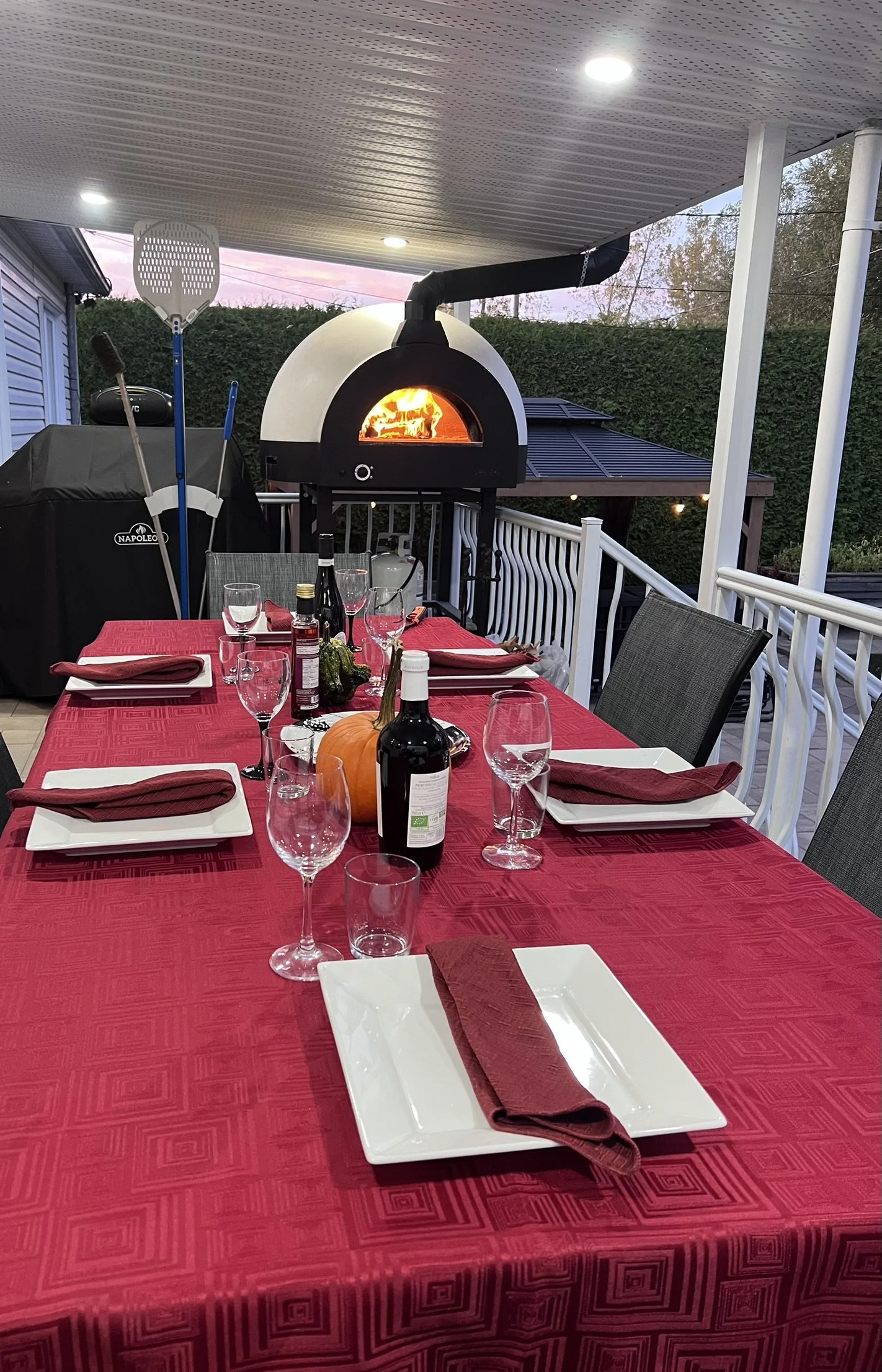 Outdoor dining table set with red tablecloth, white plates, red cloth napkins, wine glasses, a pumpkin, and bottles of wine, situated on a porch with a pizza oven in the background.