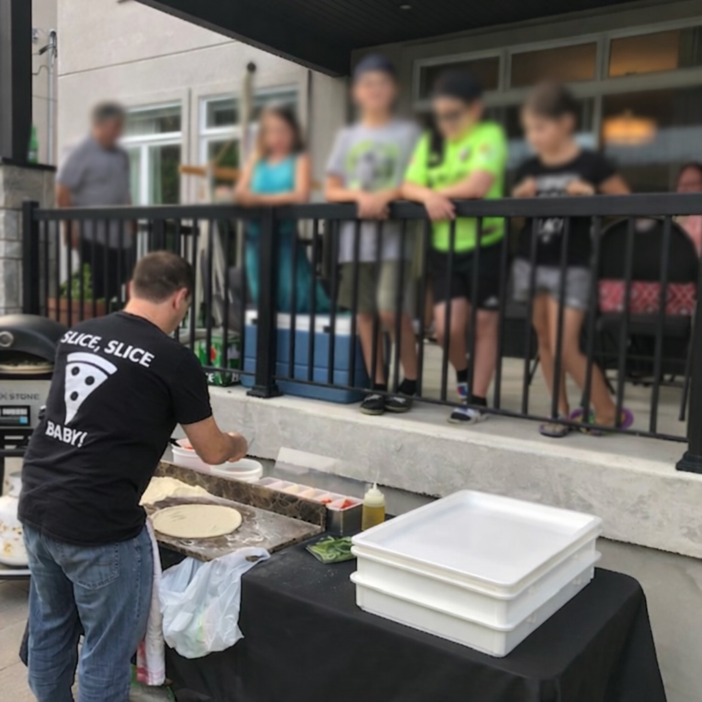 A man preparing pizza dough on a table outdoors, with kids watching behind a black fence, and a building in the background.